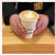 Two Hand hold a to go cup of coffee, with a latte and a flower sequence latte art made with the frothed milk. the to go cup is white with a cozy reading bread milk and honey, obscured by hand, around it. the cup is placed on a light coloured wood counter.