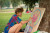 A woman is sitting in the grass at a lethbridge park [ainting on a wooden board. The woman is wearing a loose Hawaiian shirt and blue shorts, while painting a cosmic looking cat, in a bright pinkish red and blue and green rings around its head. the woman has flower earrings and has the board she is painting on leaning up against a tree. at the bottom the boar, her photos of reference can bee seen positioned for easy viewing while the woman paints.