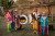 Pictured are four people in traditional Blackfoot regalia in the Fort Whoop Up interpretive centre. from left to right, there is a woman in salmon coloured dance garment, with long teal and neon green tassels and a beautiful patterned design. the woman beside her, is wearing another classic dance garment, lined with jingles and a western hat. to her right is an older gentlemen in another traditional dance garment, with feathers strewn from his hair, and feathers at both his back and in his hand, to his right is a young boy, wearing a similar dance garment to the first, but this Tim a mostly teal colour, with red white and blue tassels. behind them is a sign that reads "OKI" the Blackfoot creating. Behind them is the replica of the fort, as well as the coulees behind the fort and a tree.