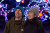 A couple is looking around themselves and smiling at the the Nikka Yuko Japanese Garden Winter Light Festival. Blurred in the background you can see all the lights of the beautiful light show in the garden. the man is wearing a teal toque and a olive green jacket. the woman is wearing a grey toque with a tan coloured pom pom and her long black hair coming out of the bottom of the toque. on top and a black jacket