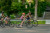 a family of four is biking in a neighbourhood road lined with trees and houses. the family is m moving past everything at a pace that leave the world around them tilting into a blur. Man leading the family with his daughter is wearing a dark brown shirt with navy blue shorts and is riding an olive green bike. the child is wearing a multi colour tank top and grey shorts, she is riding a grey bike with pink lettering. The child behind the daughter, the son, is wearing a grey shirt with beige shorts. the son is riding a bike that is black with grey and red details. behind all of them is the mother who is wearing a tan coloured shirt and grey shorts. the mother is also wearing a blue backpack.