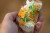 a hand is holding a small piece of bread with a white spread, orange jam and micro greens on top. the background is completely blurred.