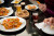 A glass topped table is sitting with plates of food, beers and two sets of hands to the right of the frame.  there are nachos and dumplings visible on the plates and in the hands of the people in the frame.
