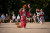 W woman in a a red kimono is in the centre of the frame and is performing a traditional dance. in her hand she has two bushles, one of auntmn leaves and one of  cherry blossoms. there is a woman behind her doing the same.