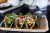 three tacos are being held and supported by a metal tray that is on a small clay plate. the tacos have corn radishes cilantro and beef in them and behind them is a small bowl with a green sauce in it. the tacos are on a black wooden table.