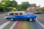 A blue car is speeding past the crowds at Lethbridge's Street Machine Weekend inLethbridge Alberta. there is a woman in the back of the car waving at the camera.