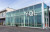 the exterior of the YQL Airport is pictured ons a sunny day, with sunlight casting a shadow on the interior. the exterior is a long rectangular glass box, with metal framing and a metal sign that says YQL on the upper right side of the front.